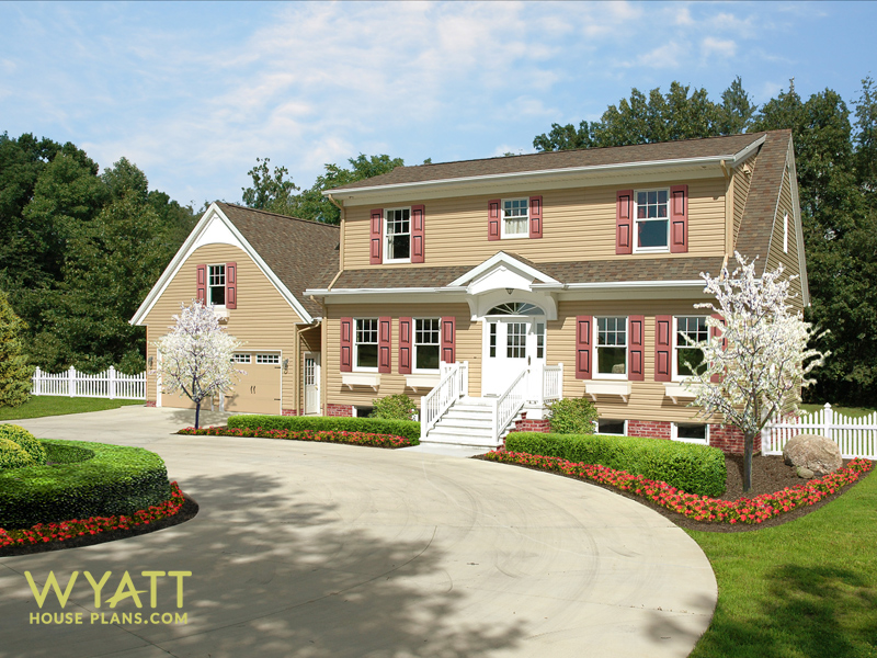 Color photo of two story home with circle drive, lots of landscape flowers and a picket fence, flowering trees, flower boxes at windows and burgundy shutters. Basement windows are above grade. An attractive home with wonderful curb appeal. Home Design by Wyatt House Plans. 3711 total square feet and up to five bedrooms. Double-hung windows. Elliptical window above entry door. Brick foundation. Bonus room above the garage.